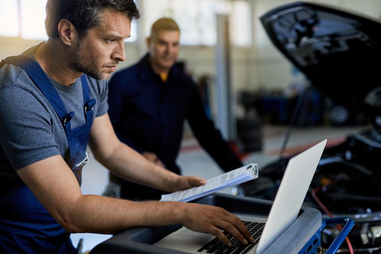 Auto repairman working on a computer while doing car diagnostic with his coworker in a workshop.